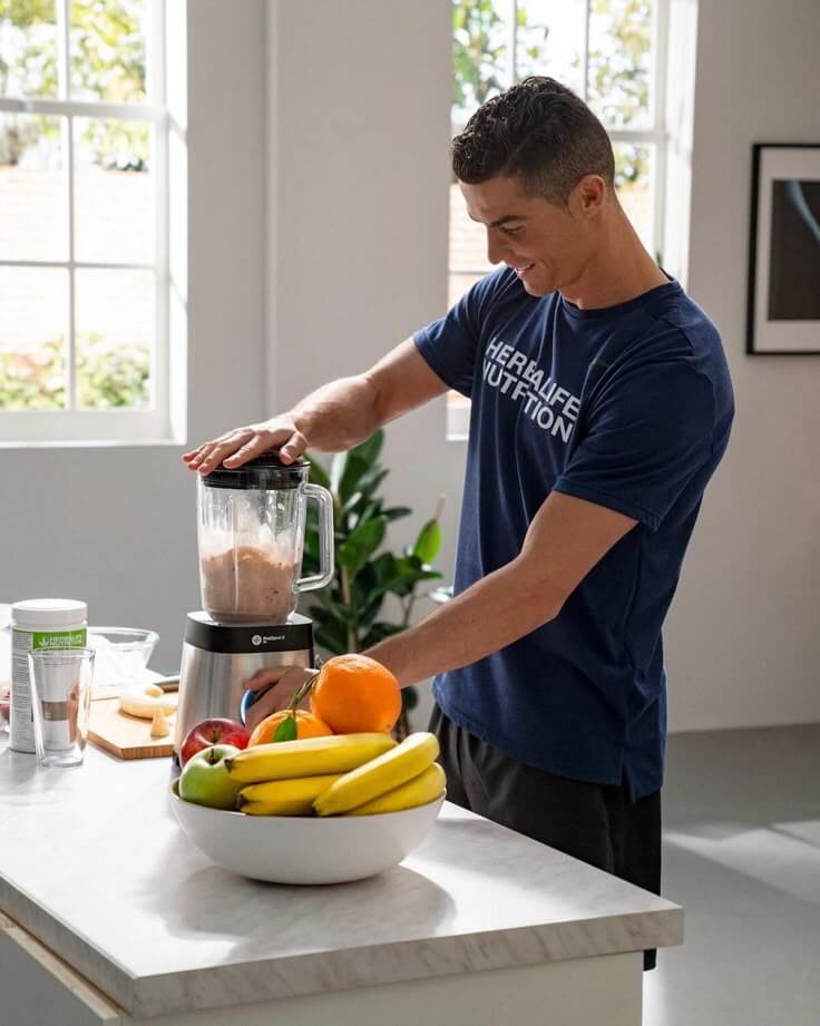 Uomo che frulla un frullato in una cucina con frutta come banane e arance sul bancone.