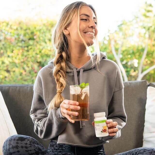 Smiling woman in a hoodie holding a refreshing drink and health supplement, enjoying a sunny day.