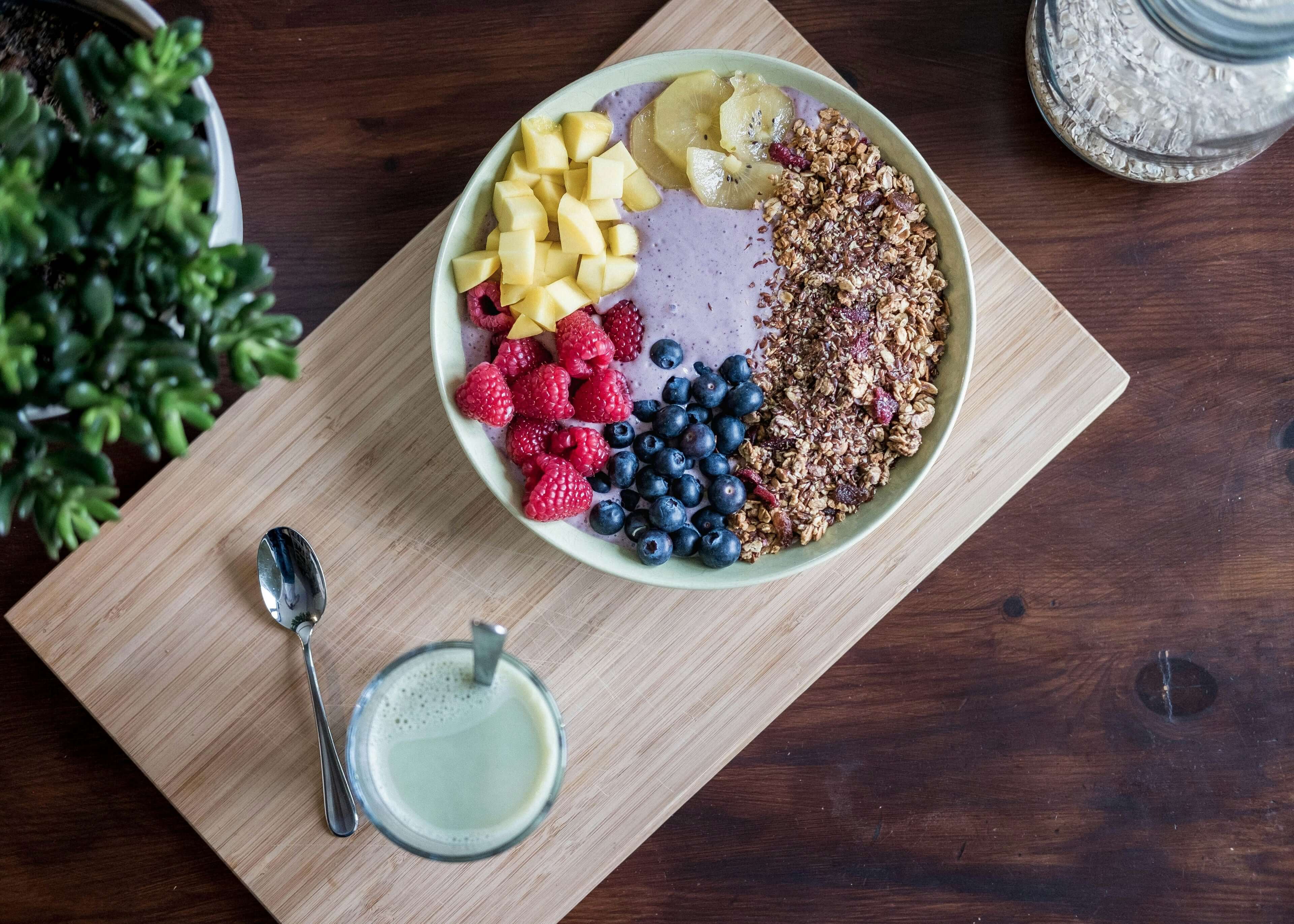 Ciotola colorata per smoothie con mango, banana, lamponi, mirtilli e granola su un tavolo di legno.