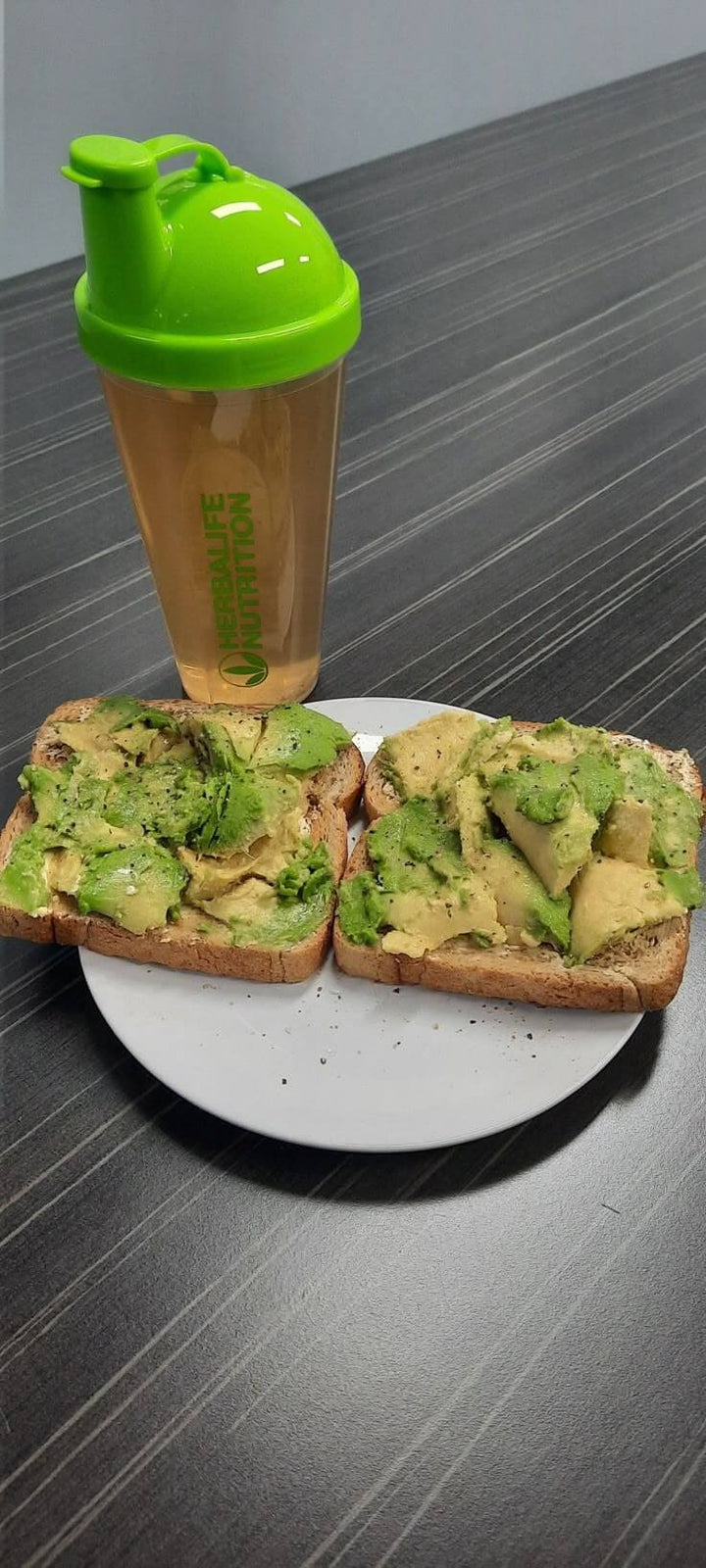 Avocado toast su pane integrale servito con uno shaker di proteine verdi su un piatto.