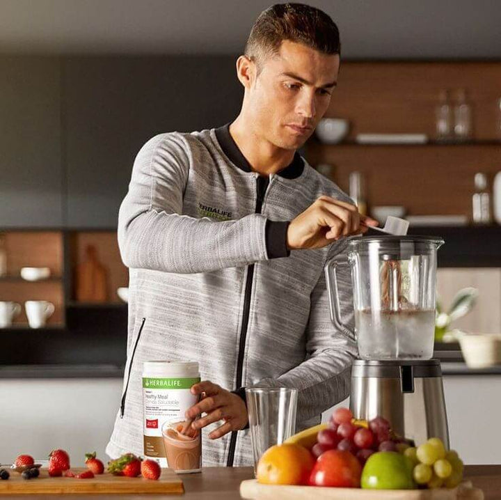 Cristiano Ronaldo prepara un frullato salutare con un frullatore e prodotti Herbalife in una cucina moderna.