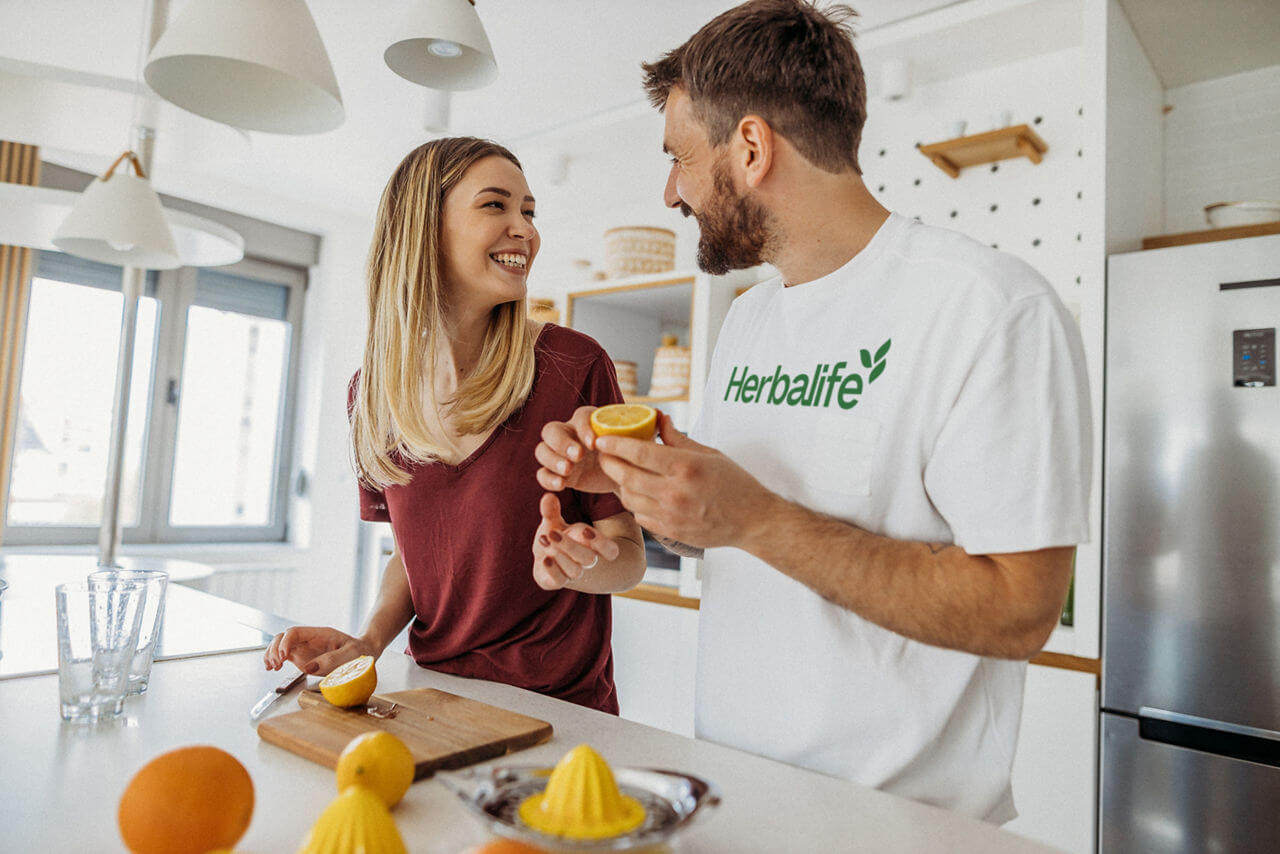 Couple preparing healthy meals in a bright kitchen, smiling and using fresh lemons with Herbalife products.